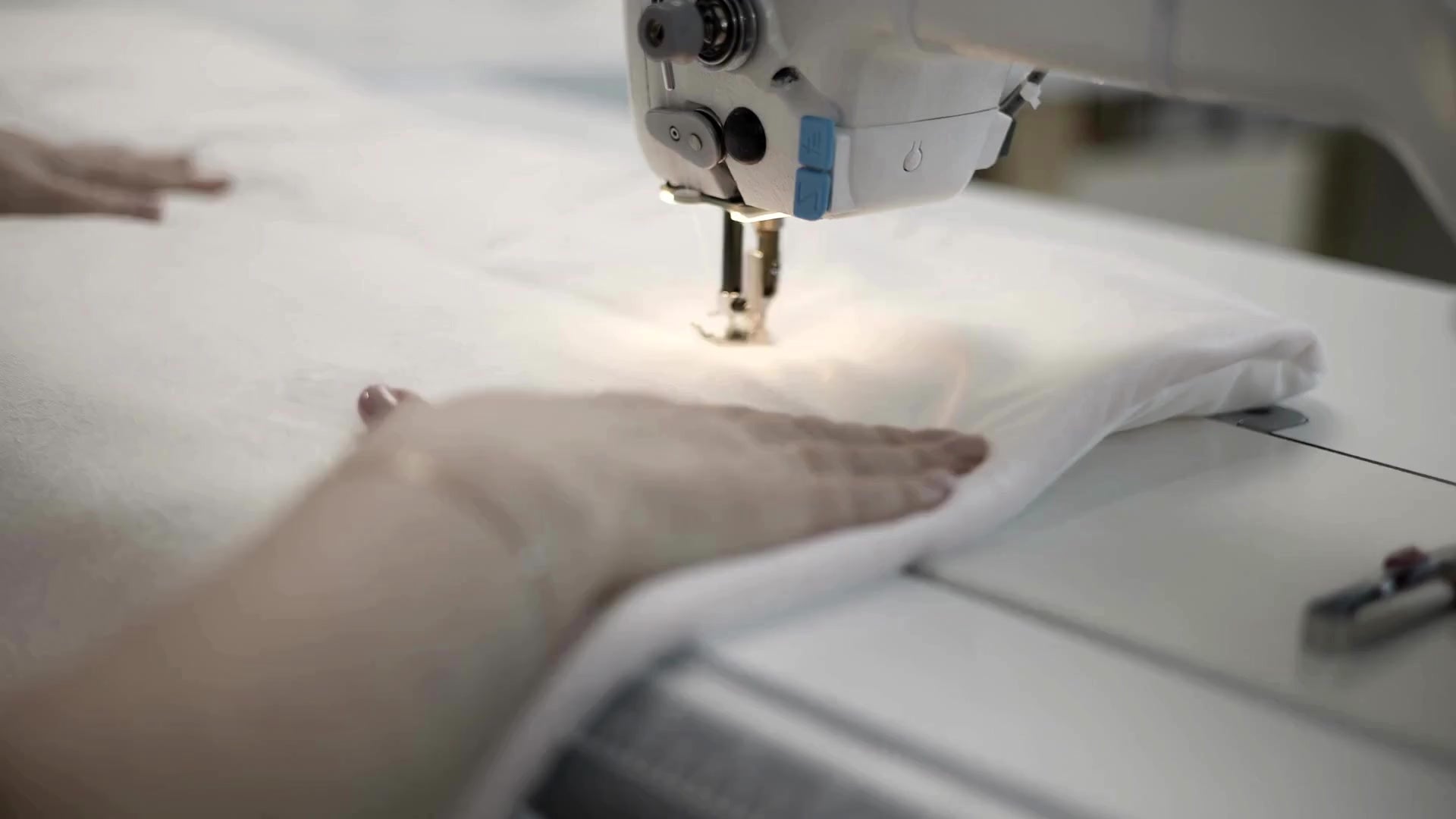Load video: Close-up of a textile worker using an industrial sewing machine to stitch white hotel linens, demonstrating precision manufacturing and quality control