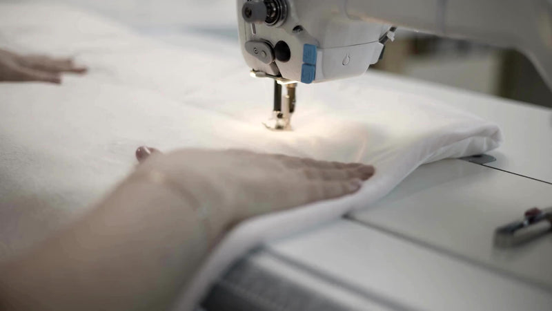 Close-up of a textile worker using an industrial sewing machine to stitch white hotel linens, demonstrating precision manufacturing and quality control